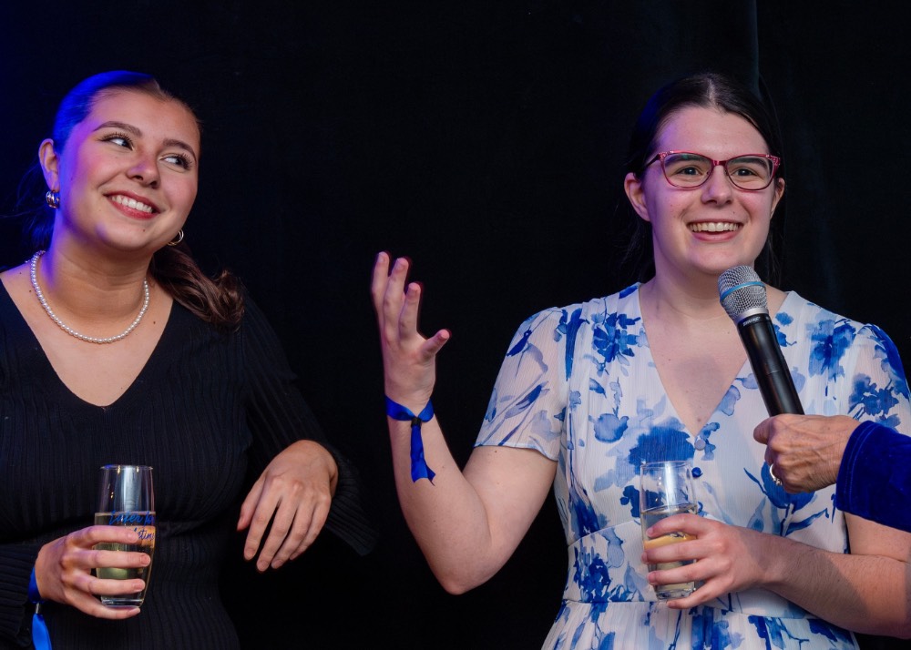 Girl smiling as she talks into microphone and the girl next to her smiles while she talks